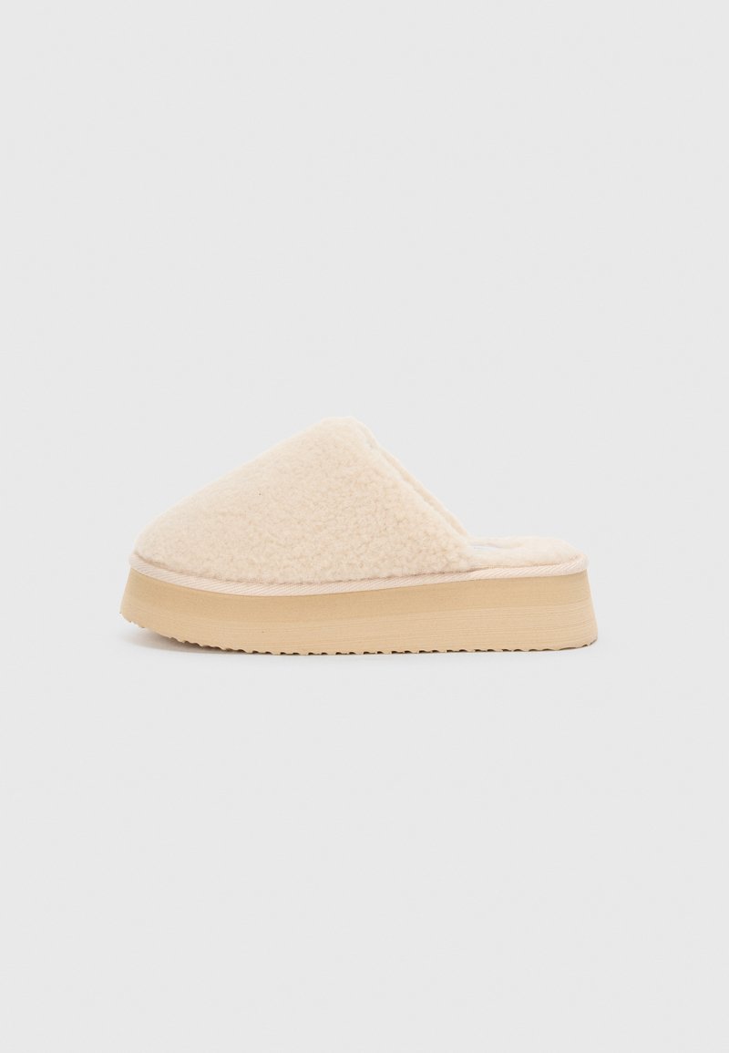 Beige plys slippers med en avrundet overdel, myk tekstur og en tykk tan plattformsåle med teksturert bunn for grep.