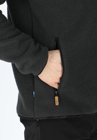 Giacca in pile nera con texture morbida, dotata di una tasca con zip sul lato sinistro e un'etichetta con logo vicino all'orlo.