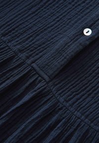 Tessuto di un blu scuro con una trama strutturata e un motivo stropicciato. Presenta una chiusura con bottone di forma arrotondata e dettagli arricciati lungo il bordo inferiore.