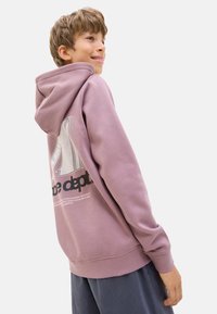 Un jeune garçon sourit en portant un sweat à capuche lavande avec un logo dans le dos, tourné légèrement par-dessus son épaule sur fond blanc.