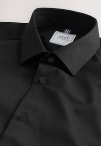 Camicia nera con bottoni, colletto strutturato, tessuto testurizzato, due bottoni neri e un'etichetta con un accento tricolore sul colletto.