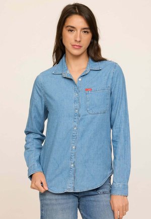 Jeune femme aux longs cheveux foncés, portant une chemise en denim bleu clair à boutons et un jean assorti, debout devant un fond uni.