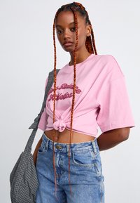 Young woman with long orange braids wearing a pink knotted crop top, high-waisted blue jeans, and carrying a gray checkered bag.