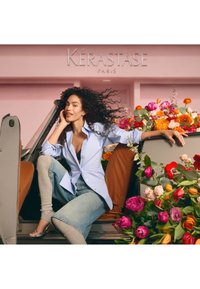 Frau mit lockigem Haar in einem blauen Hemd und Jeans sitzt in einem Auto voller bunter Blumen vor einem rosa Kérastase Paris-Hintergrund.