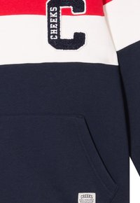 Felpa blu navy con strisce orizzontali rosse, bianche e crema, caratterizzata da una "C" ricamata e una tasca a marsupio frontale.
