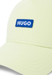 Cappello verde chiaro in cotone con visiera curvata, caratterizzato da una patch ricamata blu "HUGO" sul davanti. Texture liscia e design strutturato.
