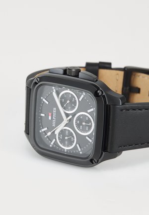Black square Tommy Hilfiger wristwatch with leather strap, three subdials showing days, 24-hour time, and date.