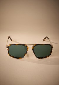 Lunettes de soleil hexagonales avec une monture écaille de tortue et des verres verts. Des accents métalliques relient les verres, et des branches fines s'étendent de la monture.