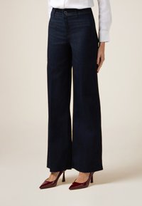 Jeans de mezclilla oscura de pierna ancha con una cintura alta, bolsillos frontales y una textura suave, combinados con zapatos de tacón alto burdeos.