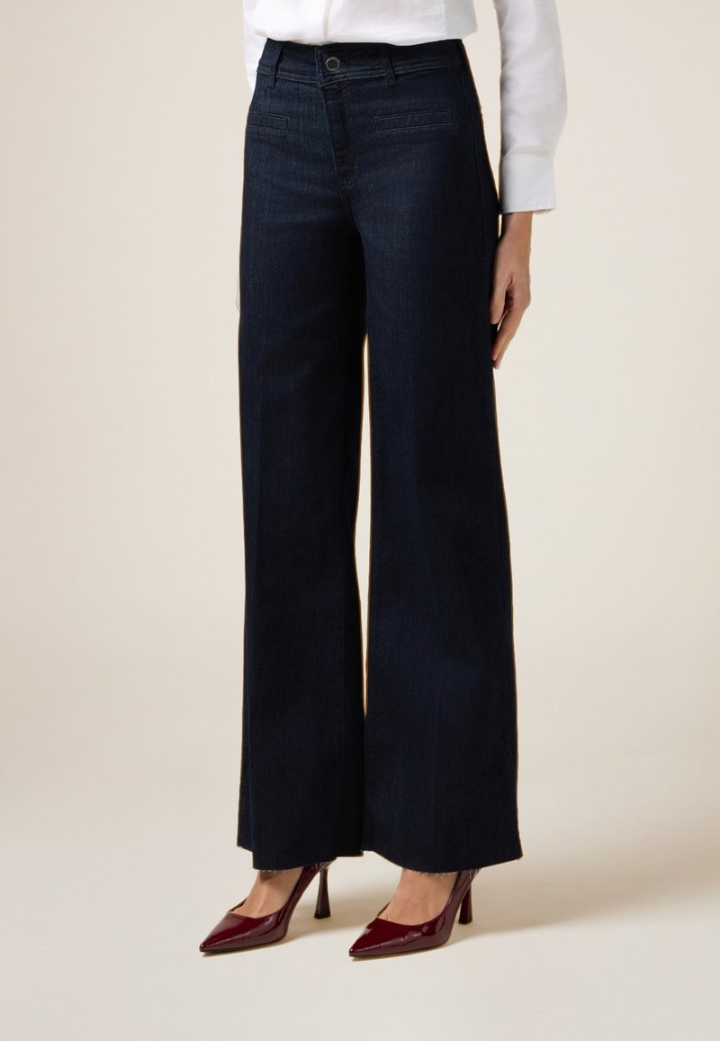 Jeans de mezclilla oscura de pierna ancha con una cintura alta, bolsillos frontales y una textura suave, combinados con zapatos de tacón alto burdeos.