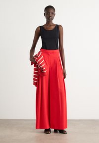 Rode hoge taille broek met plooien, gecombineerd met een zwart tanktop. Het model houdt een rood-wit gestreepte trui vast. Gladde stof, ontspannen pasvorm.