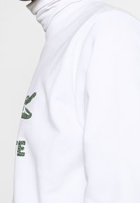 Sudadera blanca con un gráfico de cocodrilo texturizado en verde y un diseño de texto en negrita. Tejido suave con un estilo de cuello alto ajustado.
