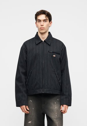 Ung mand med kort mørkt hår iført en sort pinstripe lynlåsjakke og slidte mørke jeans på en ensartet baggrund.