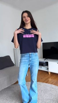 Jeune femme aux longs cheveux foncés porte un crop top bleu marine avec un texte rose et un jean bleu clair, debout dans un salon moderne.