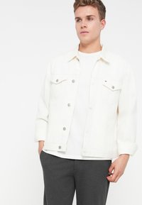 Fiatal férfi fehér farmerdzsekit visel fehér póló és sötétszürke nadrág felett, áll, kezét a zsebében, fehér háttér előtt.