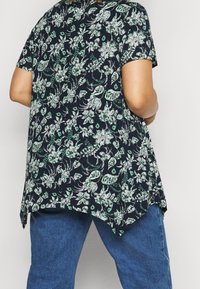 Top de manga corta con estampado floral en azul marino, con diseños en verde y blanco. Presenta un dobladillo curvado y un ajuste relajado, confeccionado en una tela ligera.