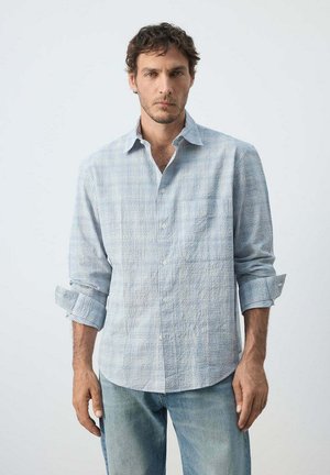 Hombre con cabello corto y rizado, vestido con una camisa de manga larga azul claro a cuadros con las mangas remangadas y vaqueros azules descoloridos, de pie frente a un fondo liso.