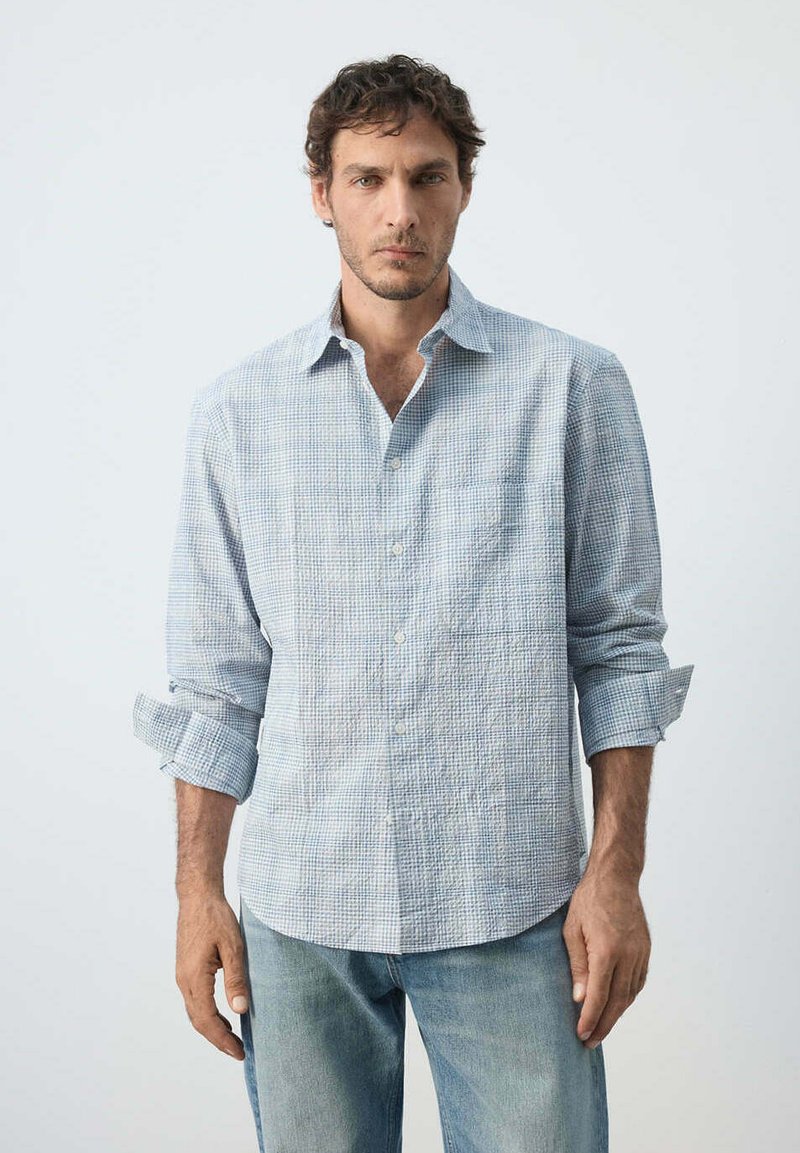Homme aux cheveux courts et bouclés portant une chemise à manches longues à carreaux bleu clair avec les manches retroussées et un jean bleu délavé, debout devant un fond uni.