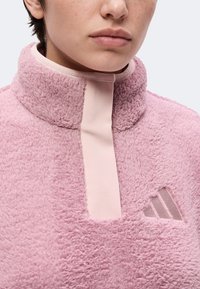 Personne portant une veste polaire rose à col montant avec une fermeture éclair verticale rose clair, arborant un logo triangulaire subtil sur la poitrine.