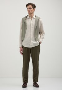 Chemise en lin beige clair avec des boutons, pantalon olive vert tapered, et un gilet en laine léger vert drapé sur les épaules. Chaussures marron.