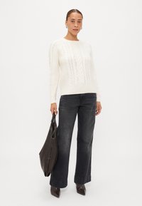 Maglione crema a maglia cable con polsini a costine, abbinato a jeans larghi scuri e stivaletti con punta a punta, mentre tiene una morbida borsa a tracolla marrone.
