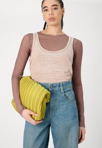 Top fără mâneci bej cu un text deschis, suprapus peste o cămașă maro cu mânecă lungă. Ținând o poșetă îngrijită, striată, de un galben pal. Blugi de culoare albastră deschisă.
