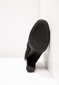 Czarny but na wysokim obcasie z wygiętym obcasem, teksturowaną cholewką i żłobioną gumową podeszwą. But jest umieszczony na białym tle.