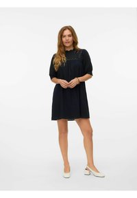 Črna obleka s kratkimi rokavi iz teksturirane tkanine in čipkastim okrasom pri ovratniku, v kombinaciji s svetlimi izreznimi čevlji.