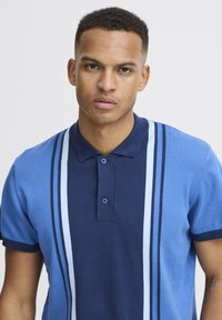 Kék és tengerészkék rövid ujjú polo póló, három gombos nyakkivágással és függőleges csíkokkal világosabb kék árnyalatokban. Sima kötött textúra.