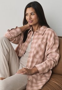 Overhemd met knopen in lichtroze ruitpatroon, gemaakt van zachte stof. Voorzien van een borstzak en opgerolde mouwen. Draagt lichtbeige broek.