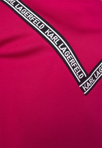 Magenta tkanina z ukośną czarną i białą taśmą z logo, na której znajduje się napis "KARL LAGERFELD", o gładkiej strukturze i minimalnym połysku.