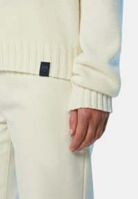Maglione lavorato a maglia color crema con polsini e orlo a costine, caratterizzato da un piccolo etichetta scura con un logo nella parte inferiore. Abbinato a pantaloni beige.