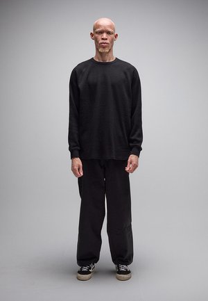 Vysoká osoba s albinizmom, oblečená v čiernej dlhoruká tričku, voľných čiernych nohaviciach a čiernych teniskách, stojaca pred jednoduchým sivým pozadím.