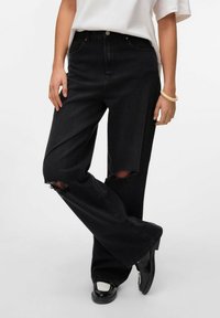 Svarta trasiga jeans med en lös passform, med slitna knän, klassisk femficksdesign och kontrasterande sömmar. I kombination med svarta skor.