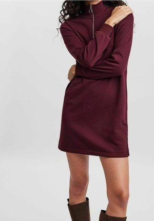 Femme portant une robe bordeaux à manches longues avec une fermeture éclair à l'avant et des bottes marron jusqu'aux genoux, debout les bras croisés.