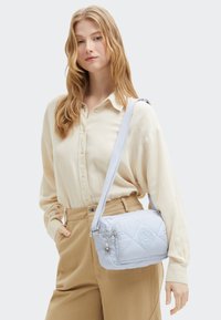 Donna bionda che indossa una camicia beige e pantaloni, con una borsa a tracolla trapuntata azzurro chiaro sulla spalla, su uno sfondo semplice.