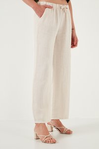 Donna che indossa pantaloni di lino beige, larghi e a vita alta con elastico in vita, e sandali beige con tacco a blocco e cinturini, in piedi con una mano in tasca.
