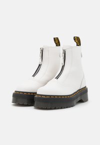 Botas de tobillo blancas hechas de cuero suave con una suela plataforma negra prominente, costuras amarillas y un cierre de cremallera lateral.