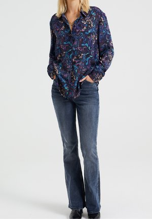 Mujer con camisa de botones azul con flores ligeramente metida en jeans bootcut desgastados, con las manos en los bolsillos, usando zapatos negros y un collar de oro.