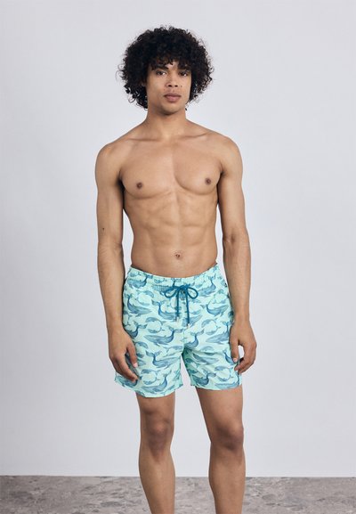 Mladý muž s kučeravými vlasmi, oblečený v svetlomodrých plavkách s tmavomodrými vzormi veľrýb, stojaci na neutrálnom pozadí.