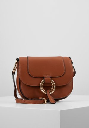 Bruine leren crossbodytas met gouden ringdetail op het voorflapje en verstelbare riem, tentoongesteld op een witte sokkel tegen een effen achtergrond.