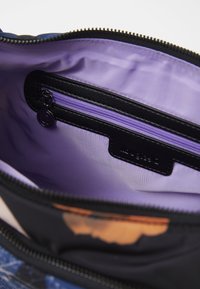 Otwarta torba pokazująca wnętrze z jasnofioletowej tkaniny oraz małą kieszonkę na zamek z czarną lamówką i okrągłym suwakiem.