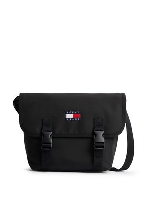 Čierna Tommy Jeans crossbody taška s klopou, dvoma prackami na zatváranie a nastaviteľným ramenným popruhom na bielom pozadí.