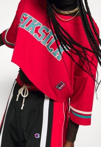 Rotes, gekürztes T-Shirt mit "SIKSILK" Grafik in Türkis, kurze Ärmel und Netzstruktur. Schwarze Hose mit weißen Streifen und Logodetails.