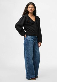 Melnas kardigāns ar teksturētu apdari, ar mežģīņu apdari ap kaklu un pogu aizdari, saskaņots ar platām zilu džinsu biksēm.