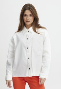 Hvid bomulds skjorte med knaplukning foran, krave og brystlomme; har en let raflet kant og sorte knapper.