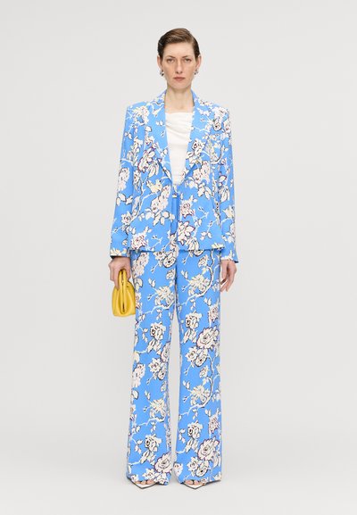Blomstret blazer og brede bukser i lyseblå med hvide og lyserøde blomster. Modellen holder en lille gul håndtaske.