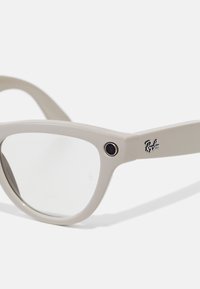 Lys beige solbriller med en rund ramme, klare linser og sorte logo detaljer på stængerne. Har et lille kamera på siden.