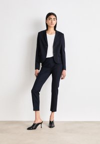 Blazer navy sartoriale con due bottoni, abbinato a pantaloni slim-fit e mules neri. La texture liscia e le linee pulite definiscono l'outfit.