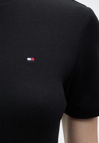 Μαύρο ριπ κοντομάνικο πουκάμισο με μικρό ορθογώνιο λογότυπο σε κόκκινο, λευκό και μπλε στο αριστερό στήθος, φορεμένο από ένα άτομο.